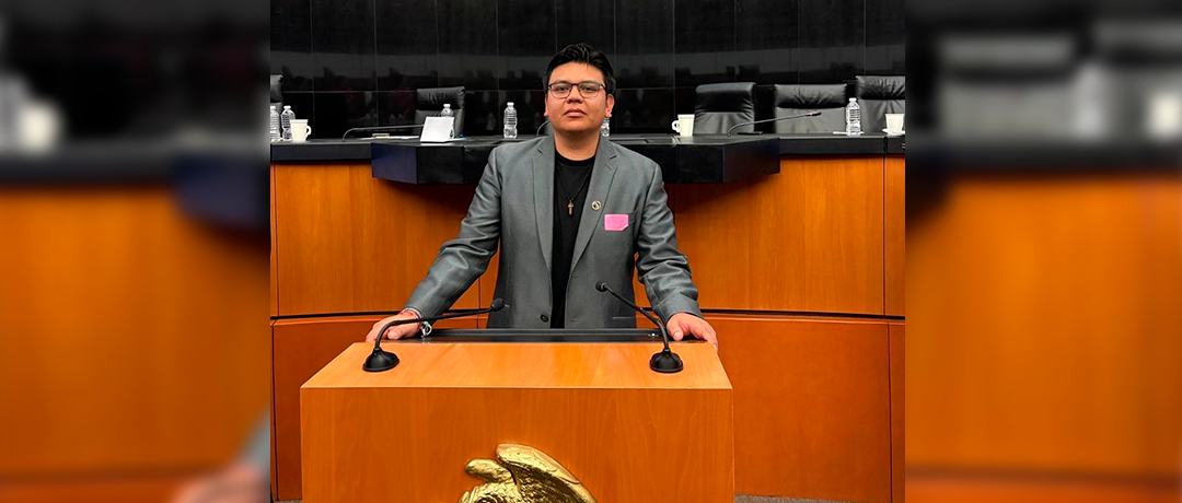 Person standing at a wooden podium in a formal chamber, wearing a gray suit and glasses, addressing an audience.