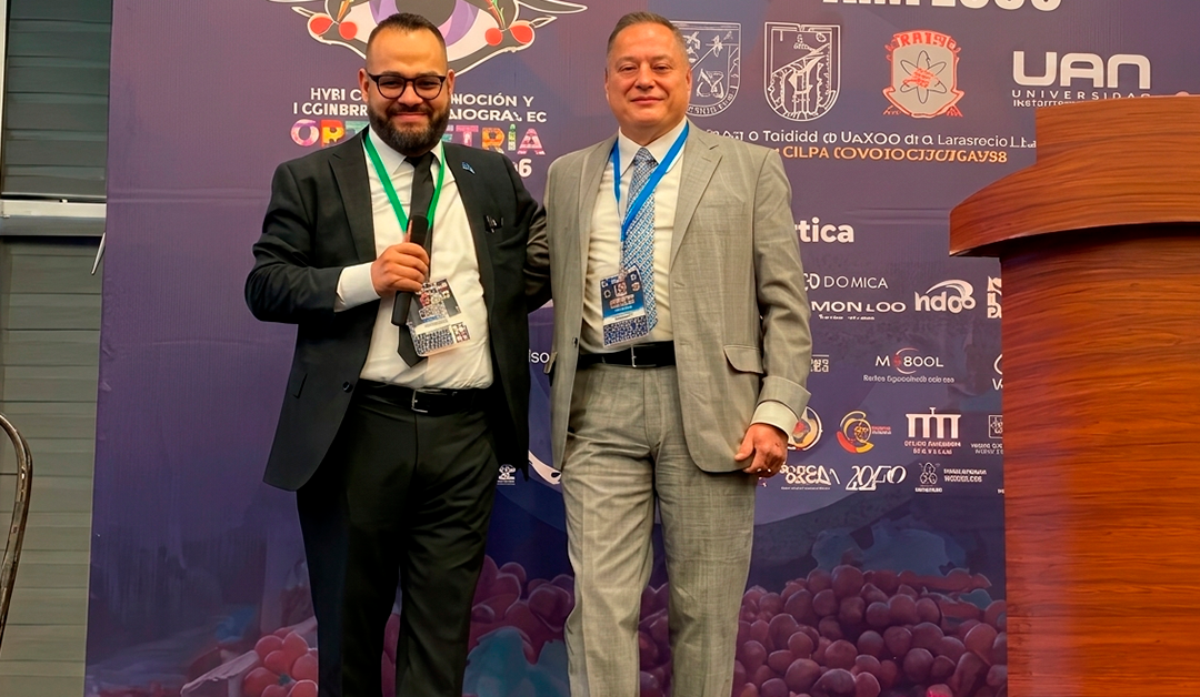 Two men in suits pose on a conference stage, one holding a microphone, in front of a logo-filled backdrop with event badges on their lanyards.