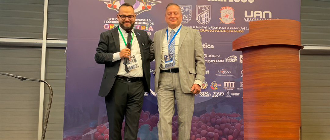 Two men in suits posing on a conference stage, one holding a microphone, with lanyards and badges, backdrop of logos behind them, podium to the right.