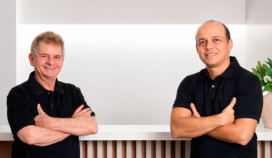 Two men with arms crossed stand at a white counter in a bright, minimalist space, wearing black polo shirts and smiling.