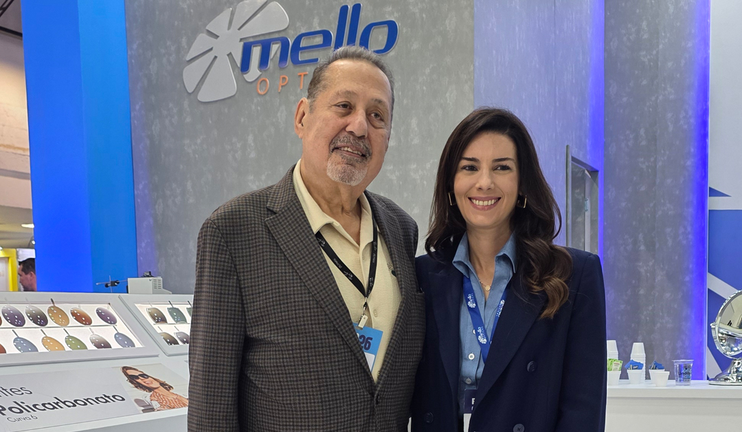 Two professionals pose and smile at a trade show booth with a Mello Optics display in the background; man in a plaid blazer and woman in a navy blazer.