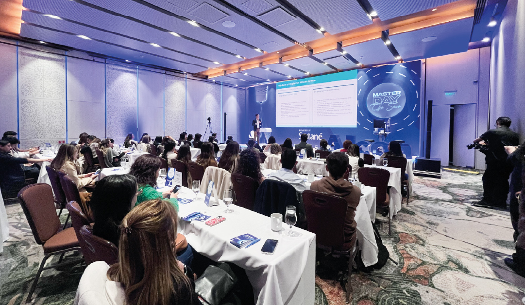 Audience seated at round tables watching a presenter on a lit stage with a large slide screen and a blue Master Day backdrop on the right side.