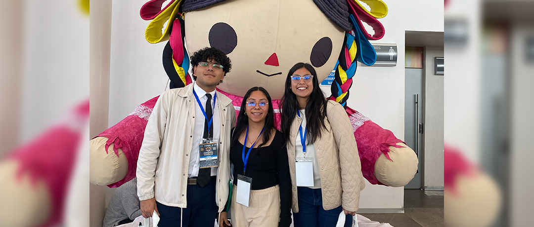 Three students with lanyards posing in front of a large colorful mascot indoors at an event.