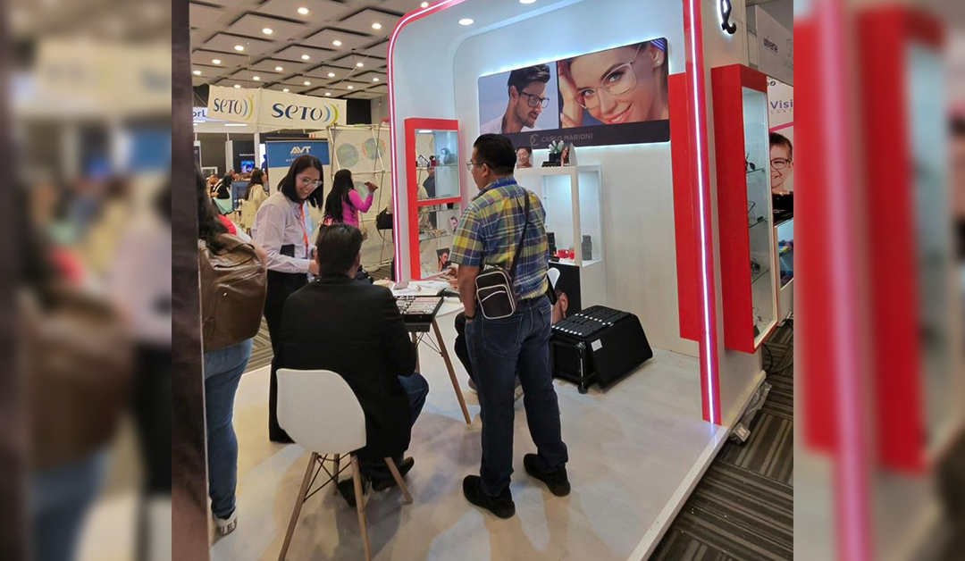 Eyewear booth at a trade show with a red and white curved display, staff talking to a visitor at a glass counter.