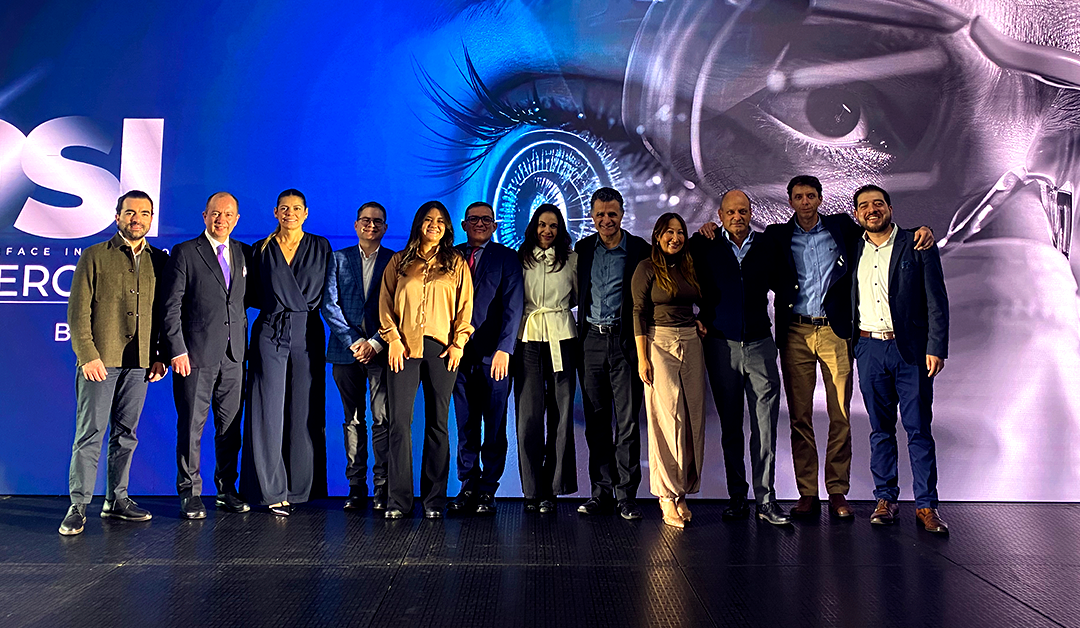 Group of twelve professionals posing on stage at a corporate event in front of a blue backdrop with an eye graphic on the left edge.