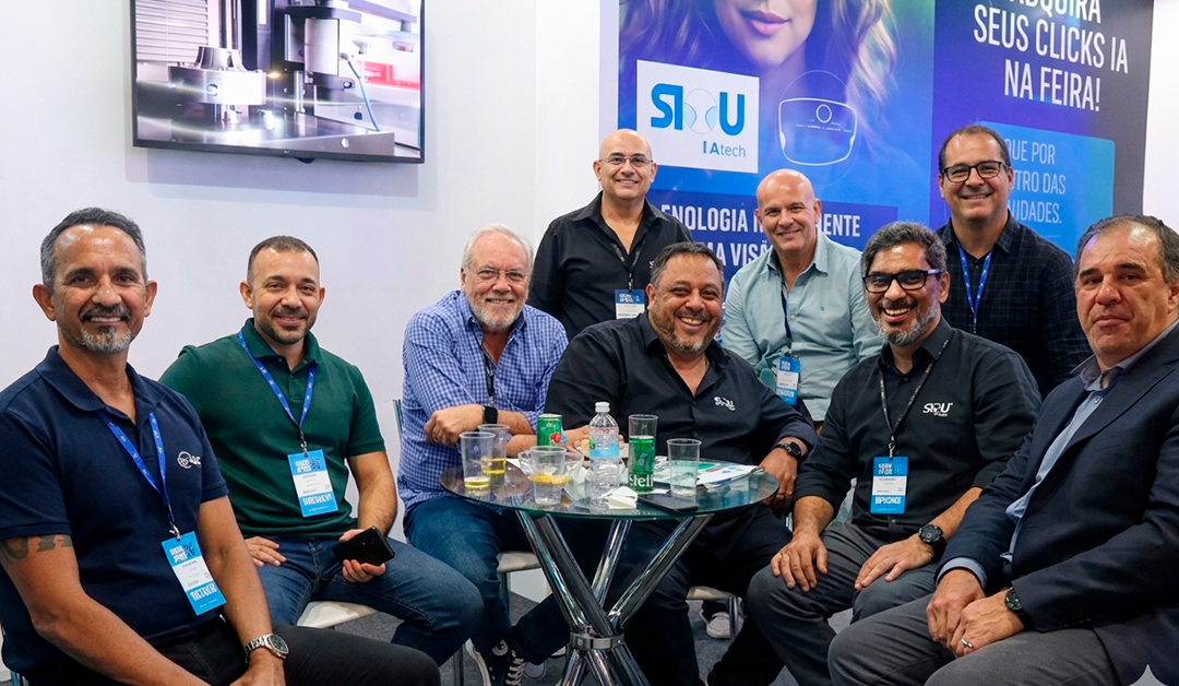 Group of men at a trade show booth, smiling and chatting around a glass table with drinks and badges visible in the foreground.