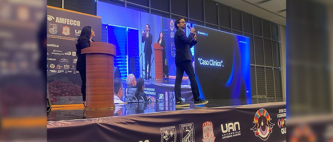 Conference stage with two podiums and presenters; a man walks across the stage while a large screen reads 'Caso Clínico'.