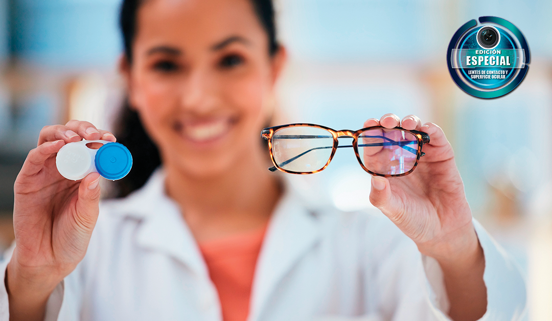 Optician in a white coat presents eyeglasses and a contact lens case, smiling at the camera, with a branded badge in the background.