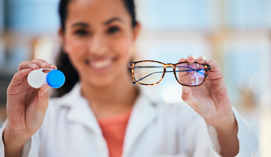 Optometrist in a white lab coat holds a contact lens case in one hand and a pair of glasses in the other, smiling at the camera.
