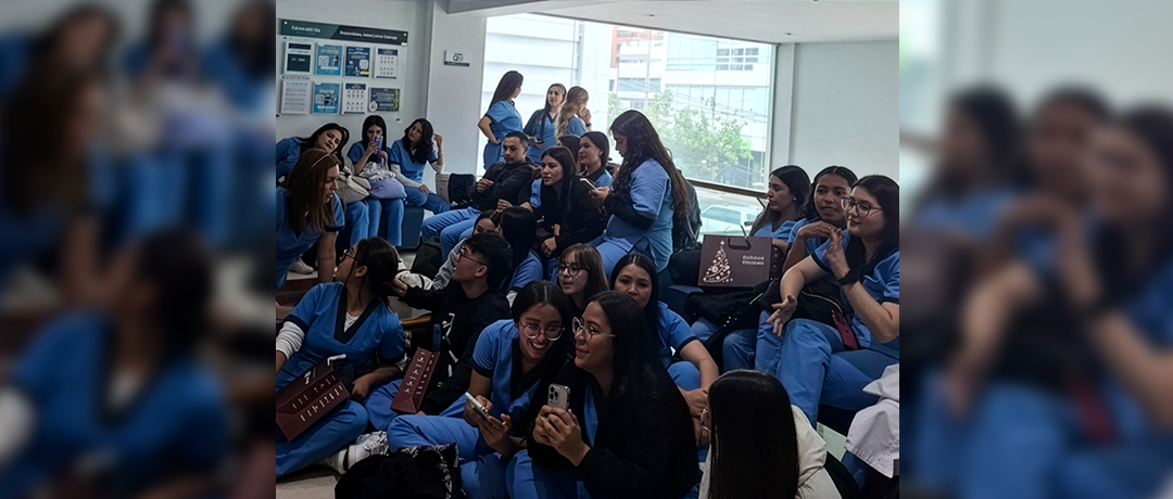 Group of nursing/medical students in blue scrubs seated in a bright lounge, chatting and using phones.