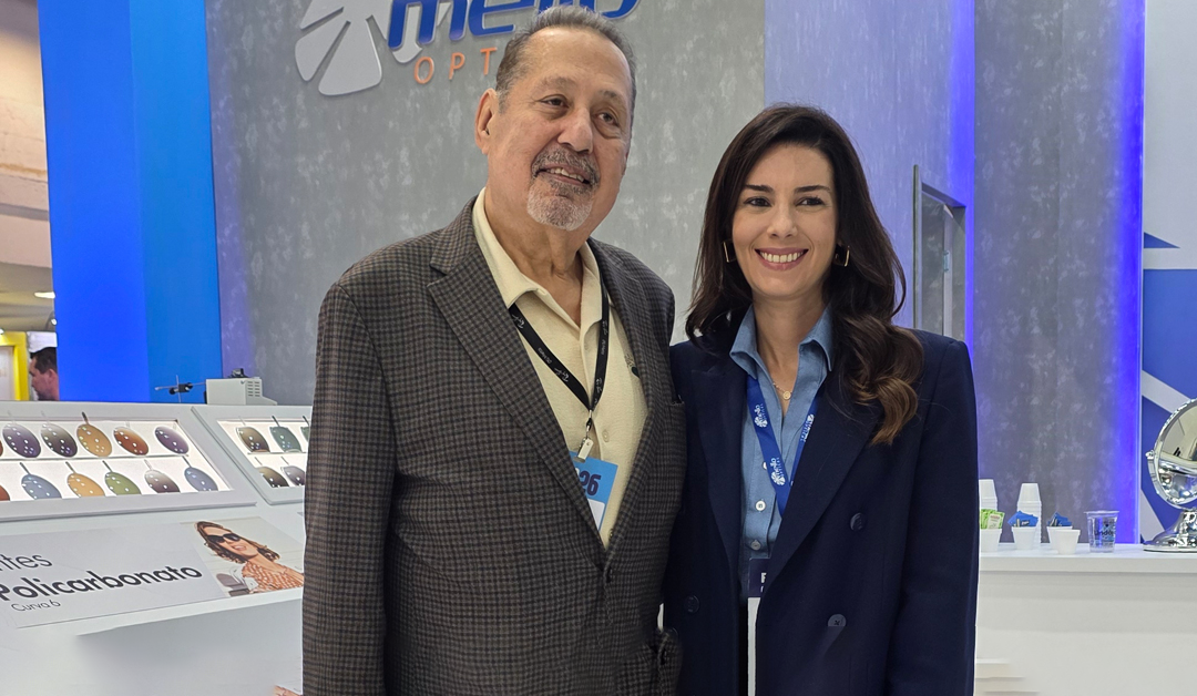 Two professionals wearing badges pose for a photo at a trade show, standing in front of a blue-lit booth with product displays in the background.