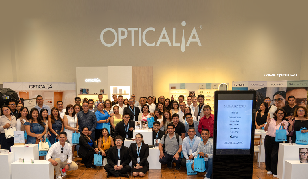 Large group of people posing at the Opticália booth during an eyewear expo, a bright wall with the OPTICALIA logo behind them? visible shelves and displays behind the crowd.