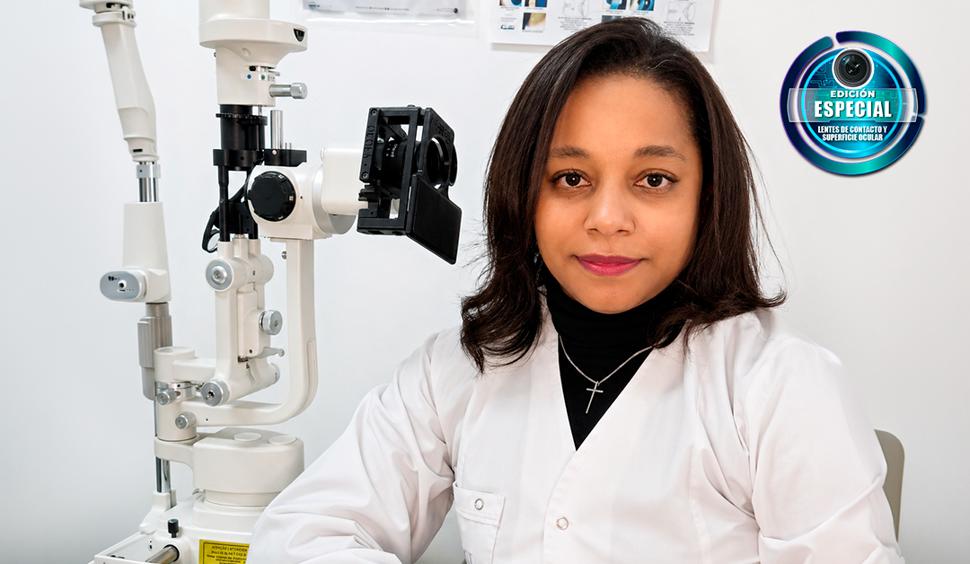 Female optometrist seated beside a slit lamp ophthalmic instrument in a clinic, wearing a white coat and black turtleneck with a cross necklace.