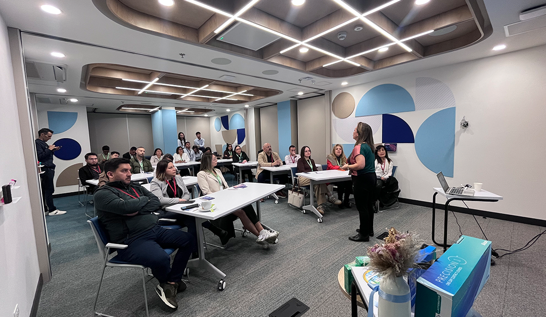 Professionals seated at tables in a modern conference room, a woman presenting at the front with a laptop and projector setup nearby.