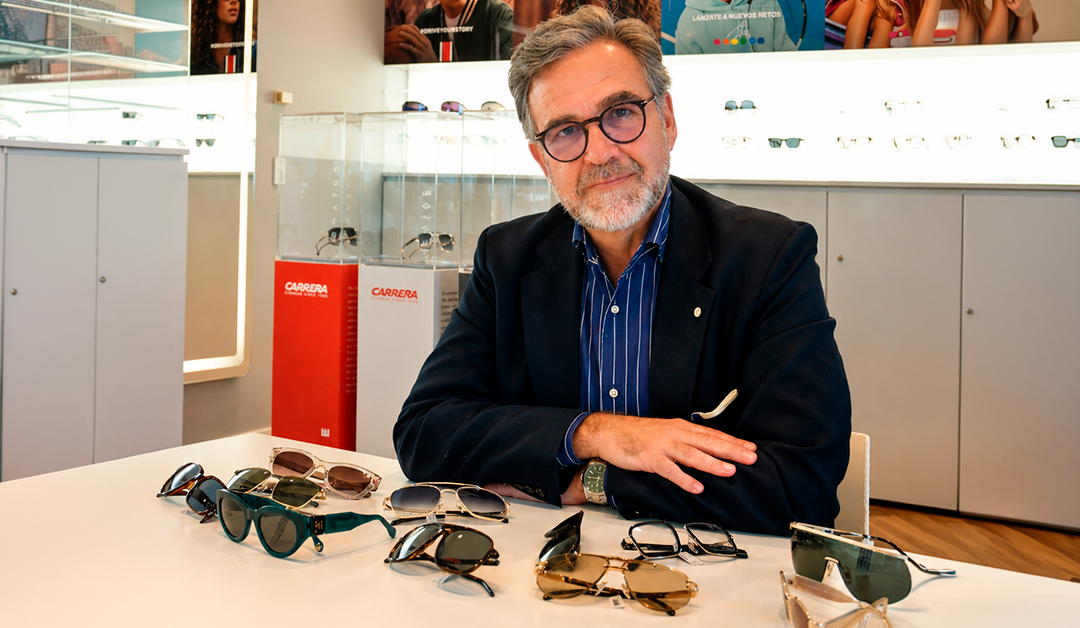 Portrait of an older man with gray beard and glasses, seated at a table covered with sunglasses in a bright eyewear store.