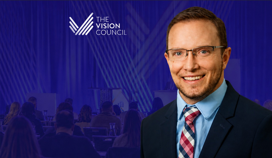 Smiling man in a suit and glasses at a conference stage with The Vision Council logo and an audience in the background.