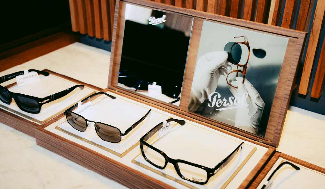 Sunglasses on a wooden display with a Persol mirror and product cards in a store window display