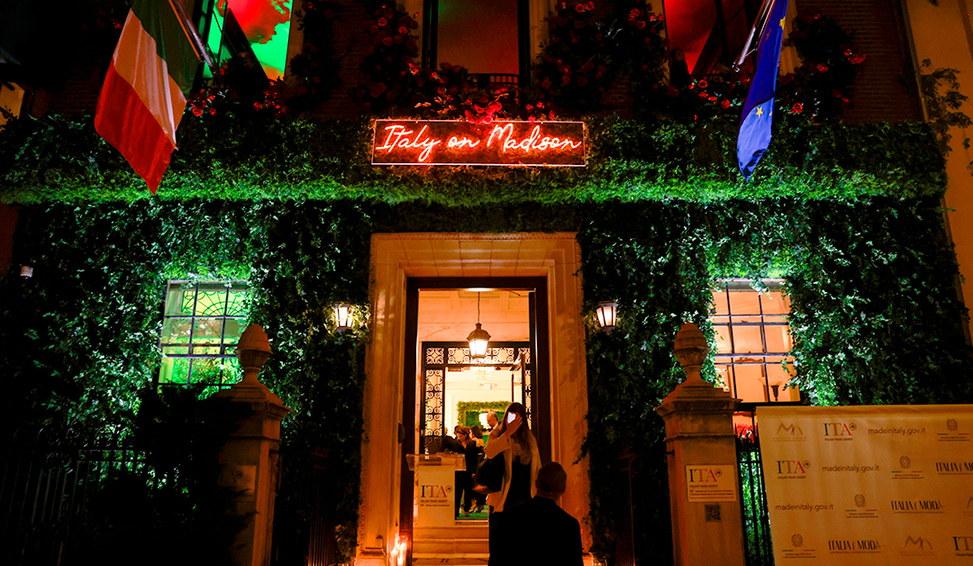 Night exterior of a greenery-covered entrance with a neon 'Italy on Madison' sign, Italian and EU flags, and guests entering the venue.