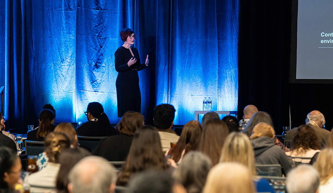 Woman in a black dress giving a talk on a stage with blue lighting to a seated audience at a conference.