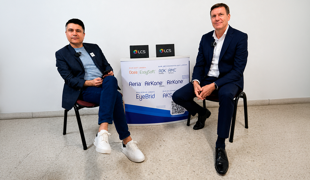 Two men in business attire sit on chairs beside a branded table displaying LCS logos and lens brand names in a bright room.