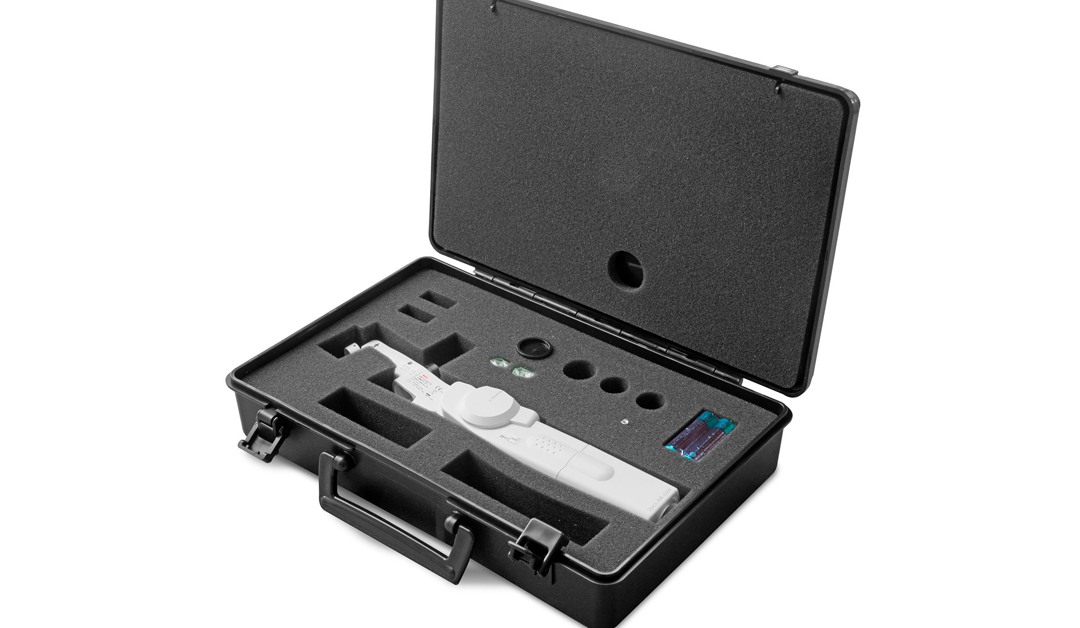 White handheld medical or laboratory device stored in a black foam-padded protective case with two batteries nearby inside the case.