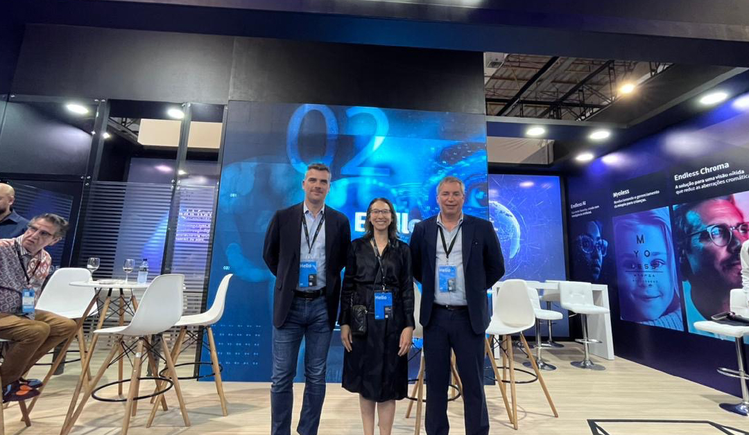 Three professionals with lanyards pose for a photo at a blue tech exhibition booth, with white chairs and high tables nearby.