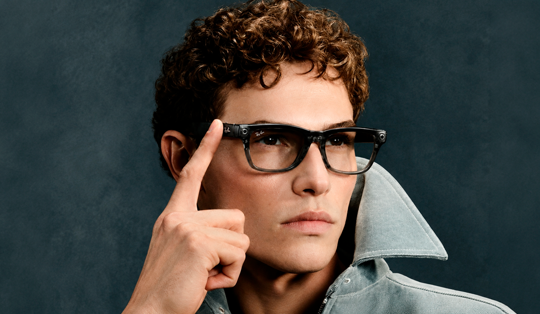 Portrait of a man with curly hair wearing black Ray-Ban glasses, touching his temple with his index finger as he gazes forward.