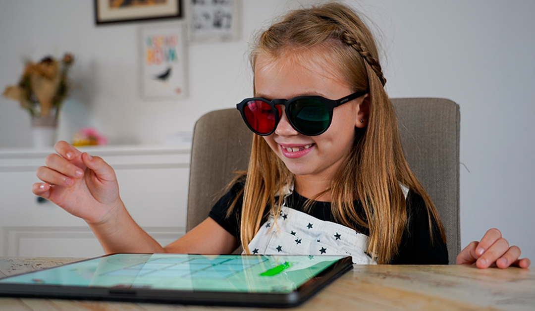Girl wearing red-tinted sunglasses taps a tablet at a table in a bright room.