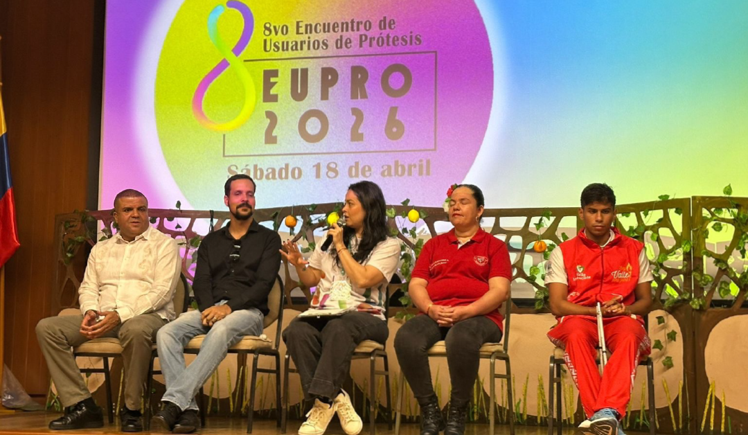 Panel de cinco personas sentadas en un escenario durante una discusión, una mujer habla con micrófono, fondo con cartel colorido que dice '8vo Encuentro de Usuarios de Prótesis EU PRO 2026'