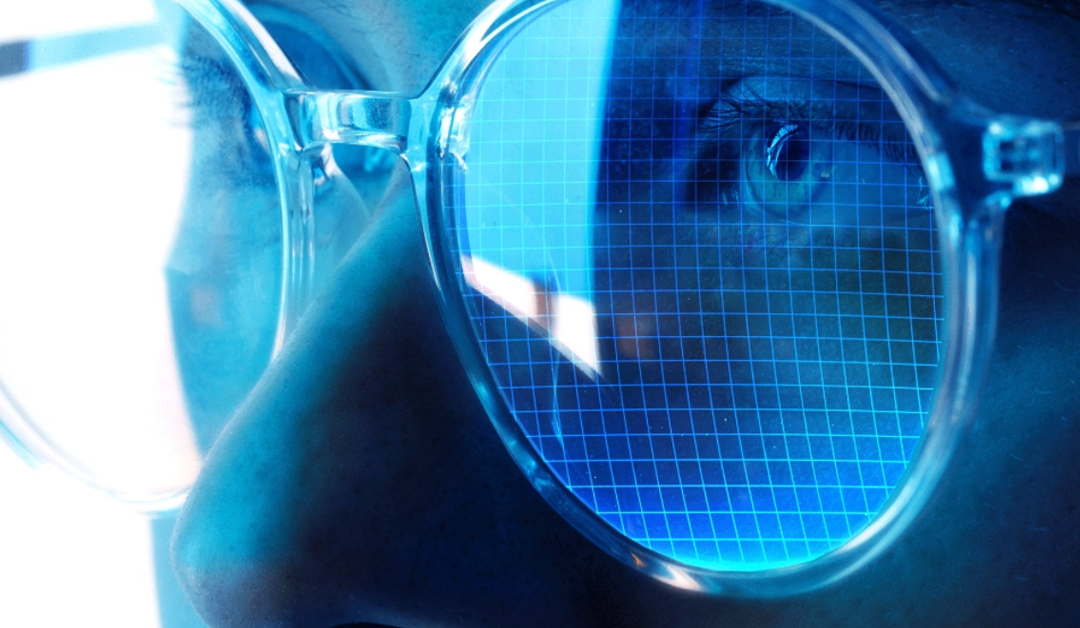 Close-up of a person wearing blue-tinted safety goggles with a grid-like reflection over the eye area, suggesting a lab setting.