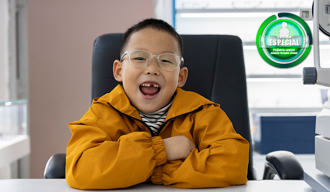 Smiling boy in an orange jacket and glasses sits at a clinic desk, arms folded, with exam equipment visible nearby.