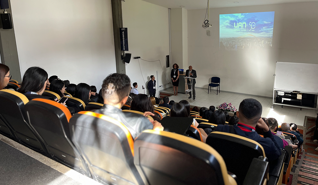 Audience seated in an auditorium watching a speaker at a podium; a projector screen shows a blue sky image and a logo on the wall towards the front of the room.
