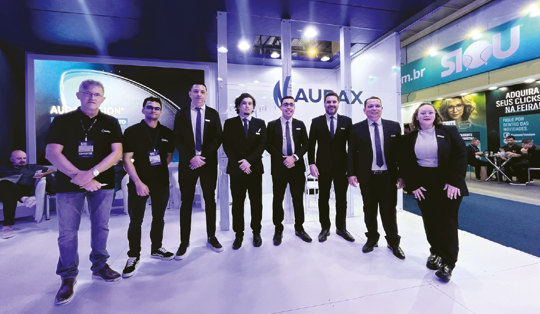 Eight professionals in suits posing for a group photo at a trade show booth, with 'AUX' branding visible in the background.