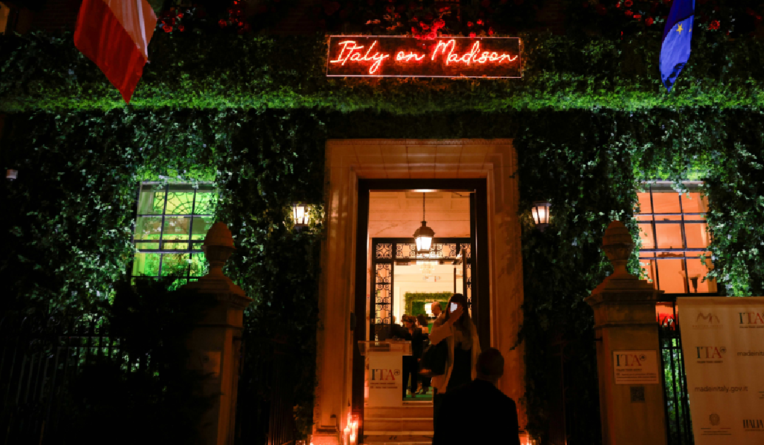 Night view of an ivy-covered building entrance with a neon sign reading 'Italy on Madison', flags overhead, and warm indoor lighting with people entering.