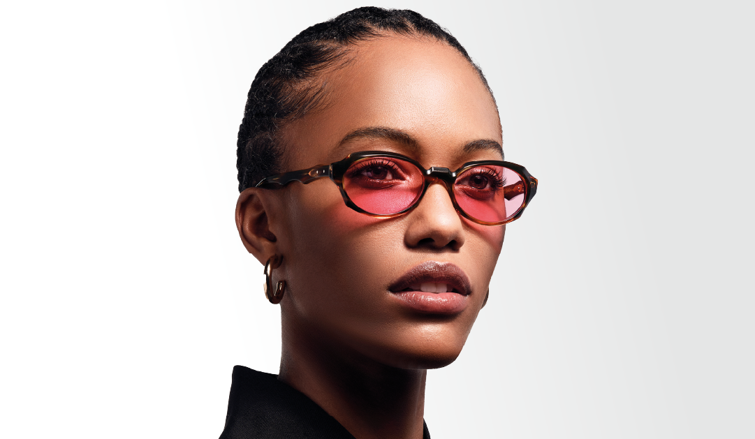 Portrait of a woman wearing pink-tinted sunglasses and gold hoop earrings against a light gradient background.