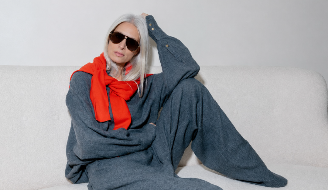 Older woman with silver hair wearing a blue knit outfit and red scarf, lounging on a light sofa in sunglasses.