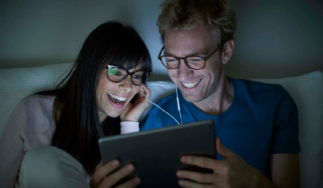 Smiling couple wearing glasses shares earbuds and watches a tablet together on a couch.