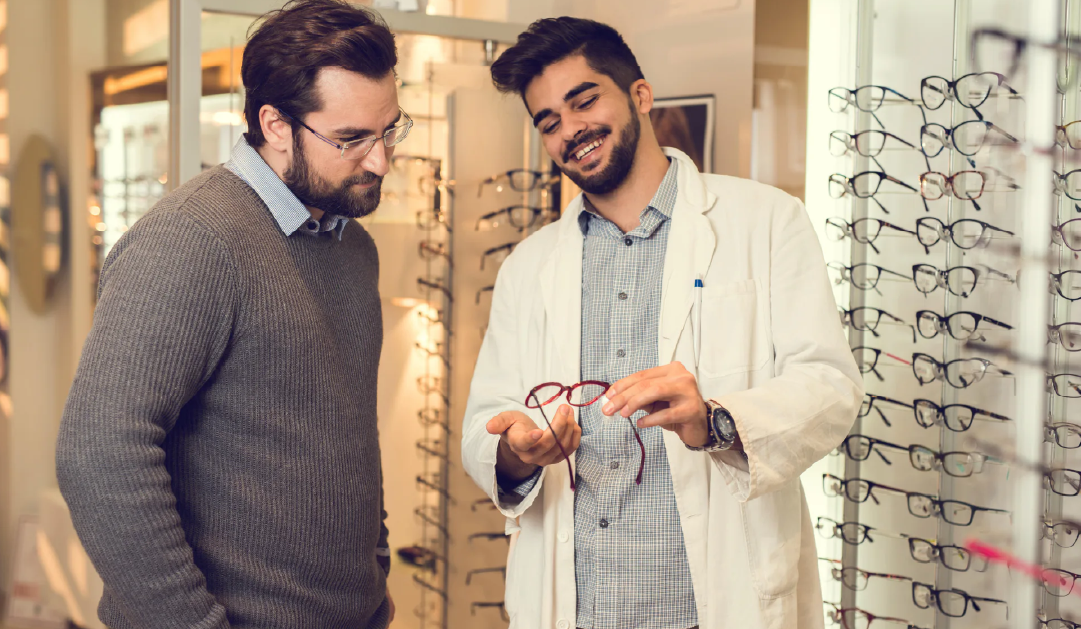 Optician in a white coat shows red-frame glasses to a male customer in an eyewear store.