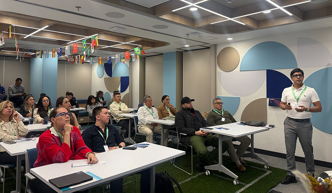 Presenter standing at the front of a classroom-style conference room, addressing a diverse audience seated at white tables.