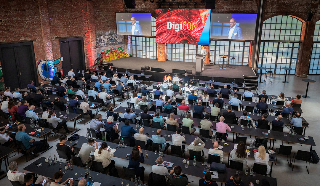 Conference hall full of attendees seated at rows of black tables, facing a stage with a podium and large DigiCON screens.
