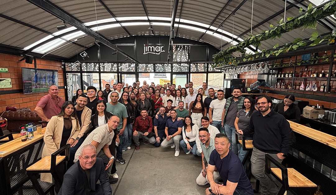 Large group of coworkers posing for a group photo in a casual, industrial-style bar/restaurant interior with a long wooden bar and exposed brick walls.