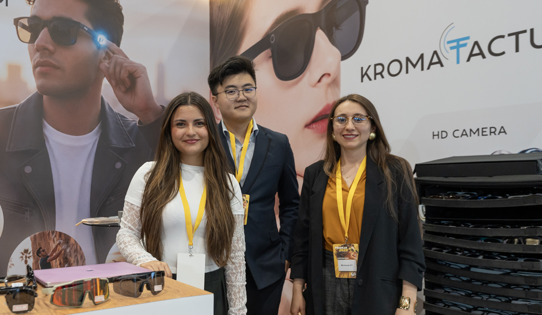 Three smiling exhibitors with yellow lanyards pose at a sunglasses booth, with eyewear on the display table in front of them and a large branded backdrop behind.