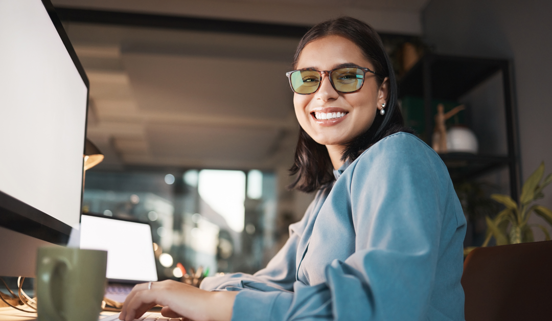 Smiling woman wearing glasses works at a desk with a computer in a modern office setting.