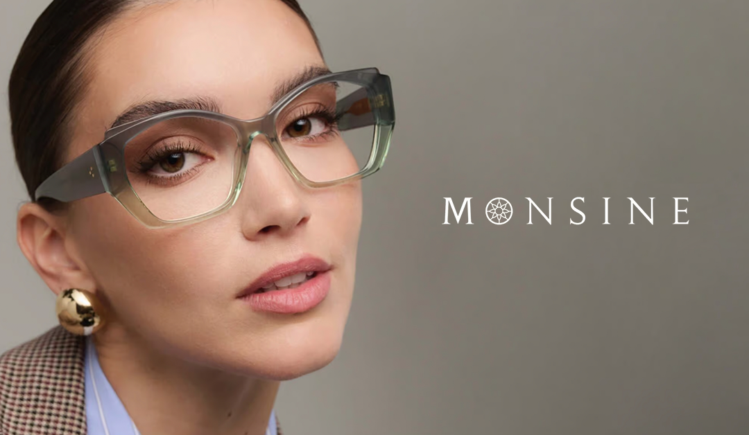 Close-up of a woman wearing oversized transparent glasses, with a gold hoop earring and a blazer, MONSINE logo on a grey background.
