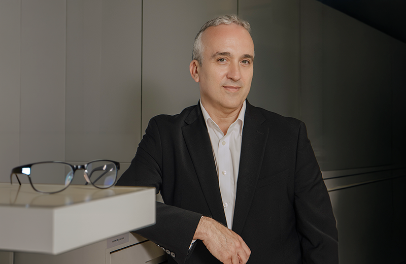 Middle-aged man in a black blazer and white shirt leaning on a white office counter with glasses nearby on the counter.