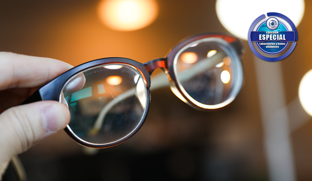 Close-up of a hand holding round glasses with reflective lenses; warm blurred background and a circular logo in the top-right corner.
