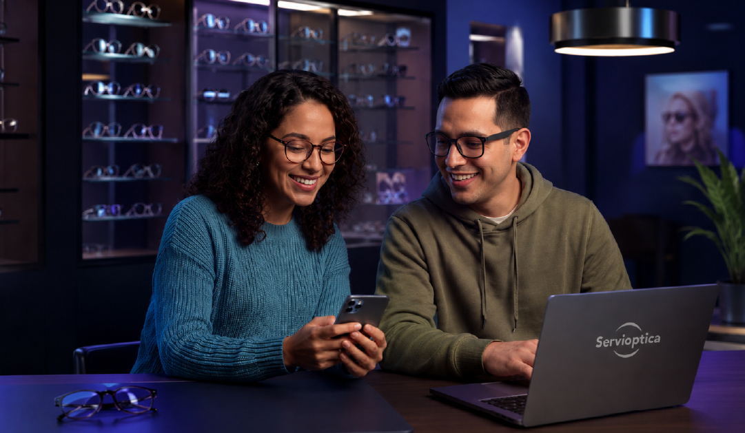Two customers wearing glasses sit at a dark table in an eyewear store, looking at a smartphone together with a laptop labeled Servioptica nearby.