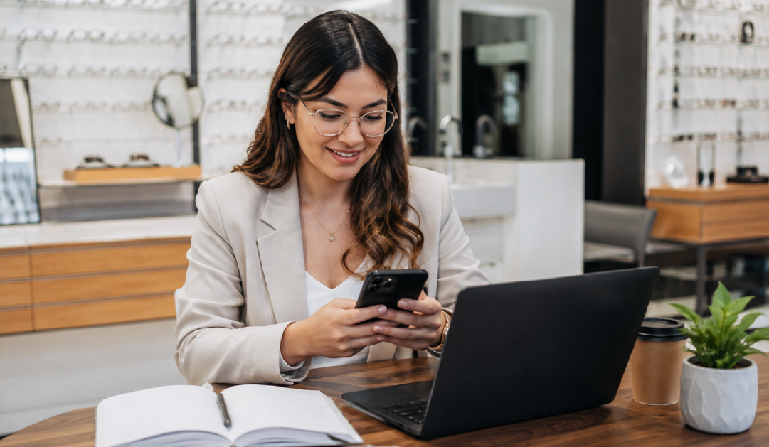 Professional woman in a beige blazer sits at a table, looking at her phone with a laptop, notebook, coffee, and a small plant nearby.