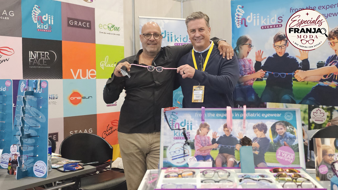 Two men pose at a colorful eyewear booth, one holding pink-rimmed glasses for a photo.