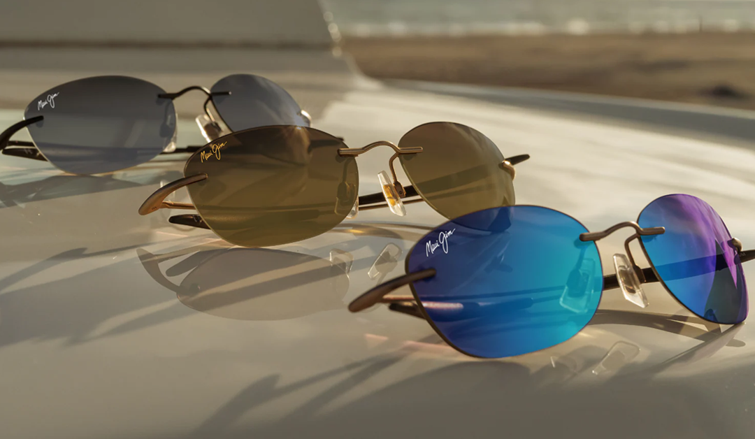 Three pairs of round sunglasses with colored lenses (blue, brown, and gray) resting on a glossy surface, with light reflections and brand logos visible on the lenses (Maui Jim).
